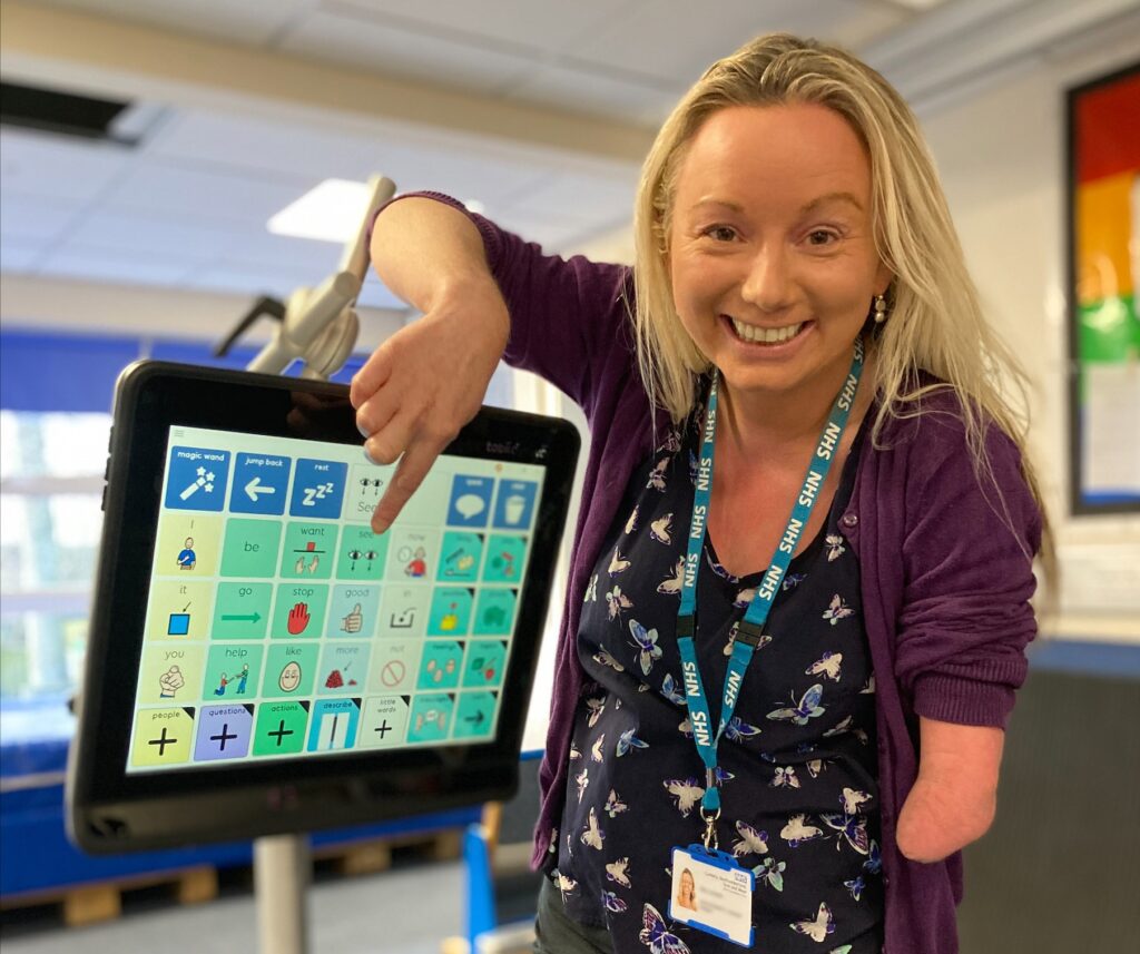 Photo of member of staff with a limb difference, pointing to an icon on a communication aid