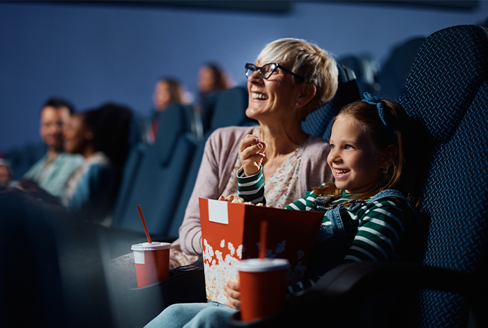A grandmother and grandchild watching a film in a cinema. Both are smiling and holding a bucket of popcorn.